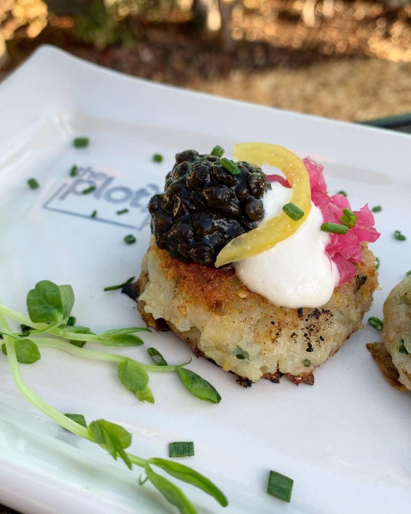 A gourmet dish featuring a potato cake topped with black caviar, sour cream, pickled onions, and a lemon slice, garnished with fresh herbs on a white plate.