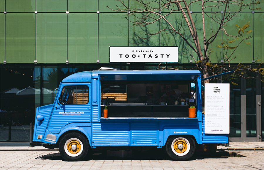 Blue food truck parked in front of a modern building with a sign reading "TOO TASTY."