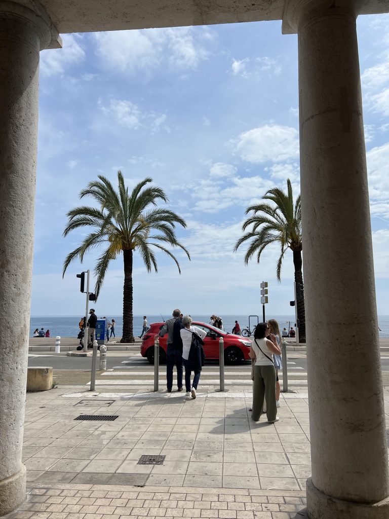 View from a columned walkway overlooking a promenade with palm trees, people, and a red car near the seaside.