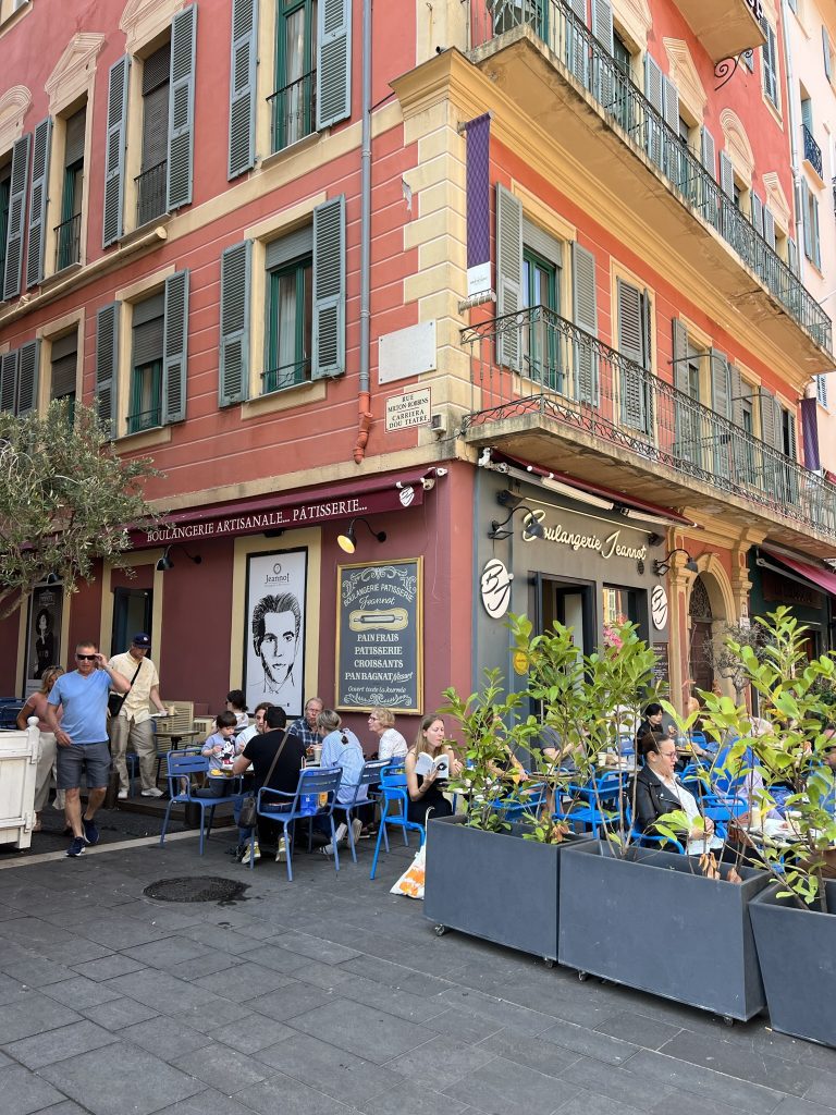 People sitting at outdoor cafe tables by a red building with green shutters and balcony.