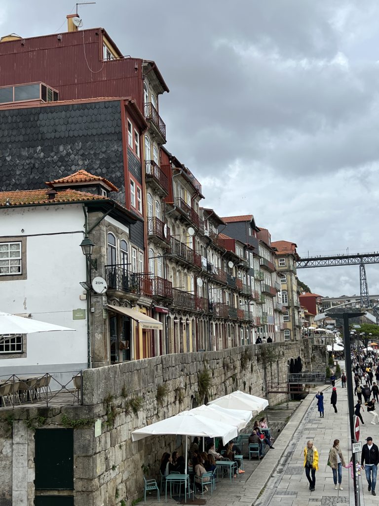 Street view with historic colorful buildings alongside a stone wall, outdoor café seating with umbrellas, and people walking under an overcast sky.