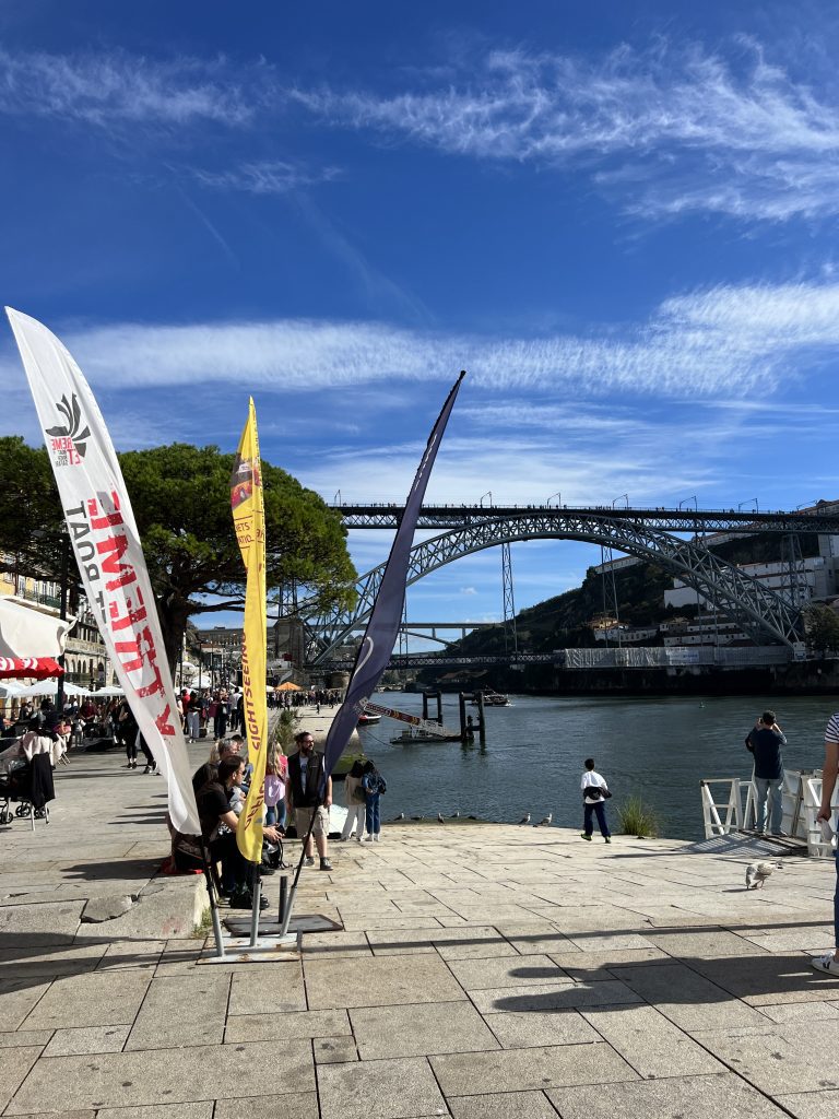 Riverfront promenade with people, colorful flags, and a bridge in the background on a sunny day.