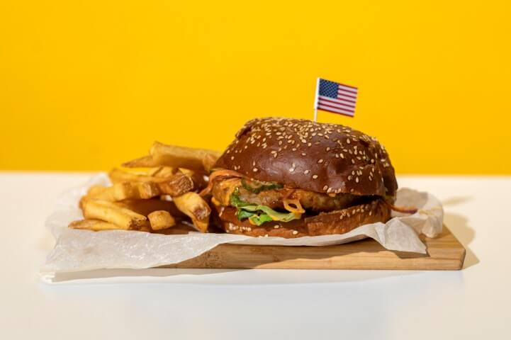 Burger with fries on a wooden board, topped with a small American flag, against a yellow background.