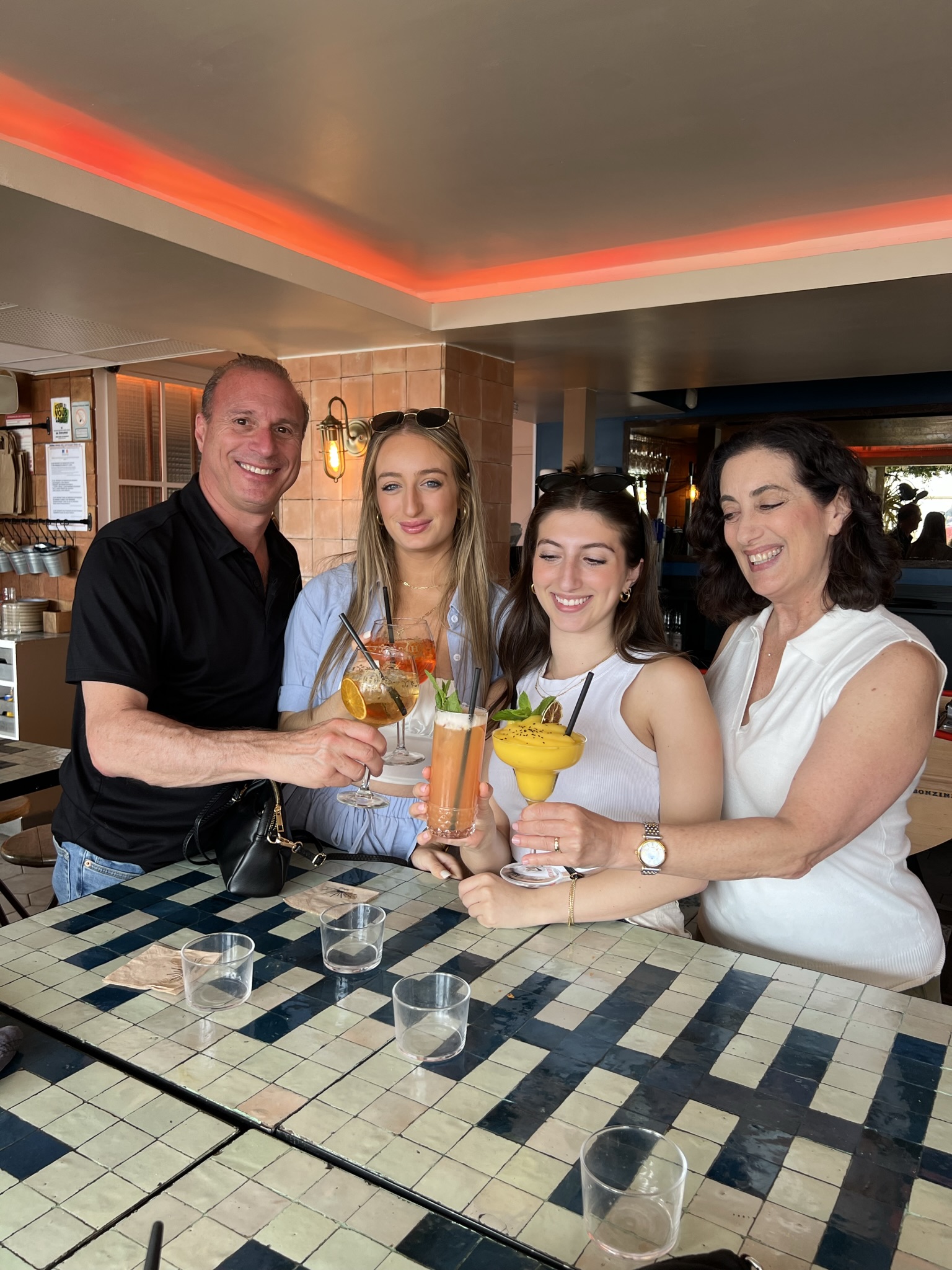Four people standing together, holding colorful cocktails in a restaurant setting, smiling at the camera.