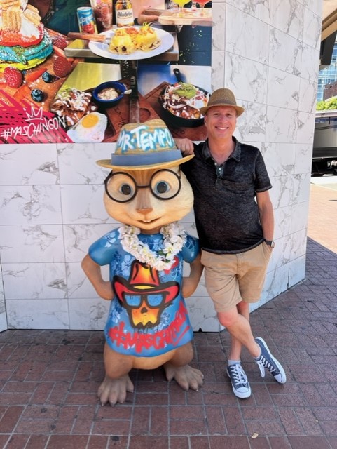 Man posing with a cartoon chipmunk statue wearing a Hawaiian shirt and hat.