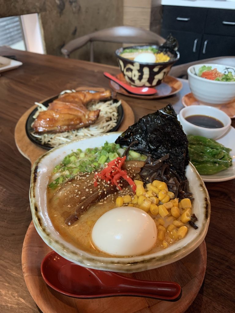 A bowl of ramen with corn, seaweed, egg, and vegetables on a wooden table, accompanied by grilled meat and a side salad.