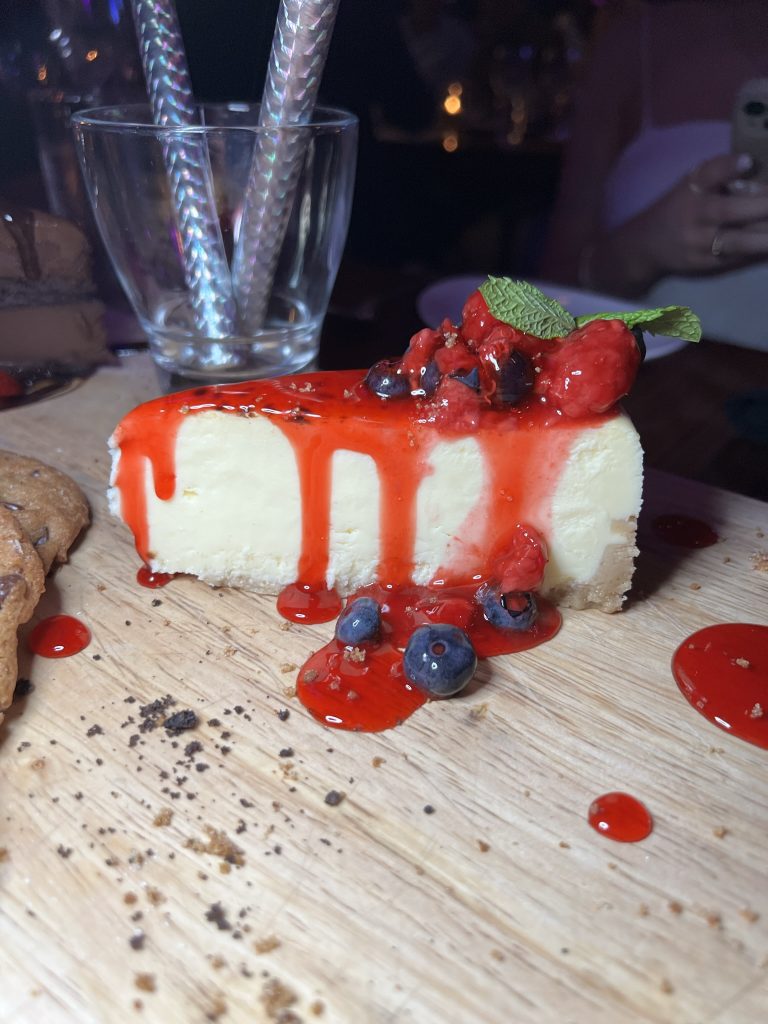A slice of cheesecake topped with red berries and sauce on a wooden board.