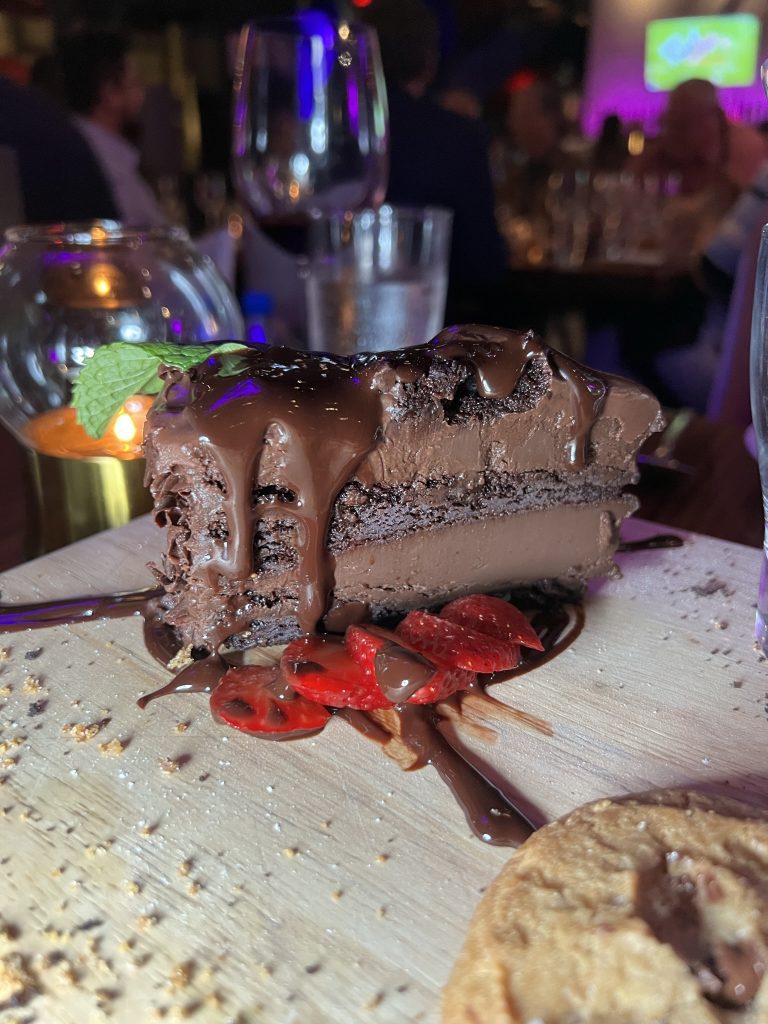 A slice of rich chocolate cake with chocolate drizzle, garnished with sliced strawberries and a fresh mint leaf, set on a wooden board beside a cookie.