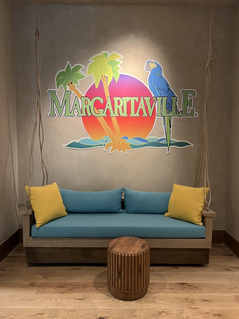 A wooden swing bench with blue and yellow cushions, a small round wooden table, and a "Margaritaville" sign featuring a parrot and palm trees on the wall behind it.