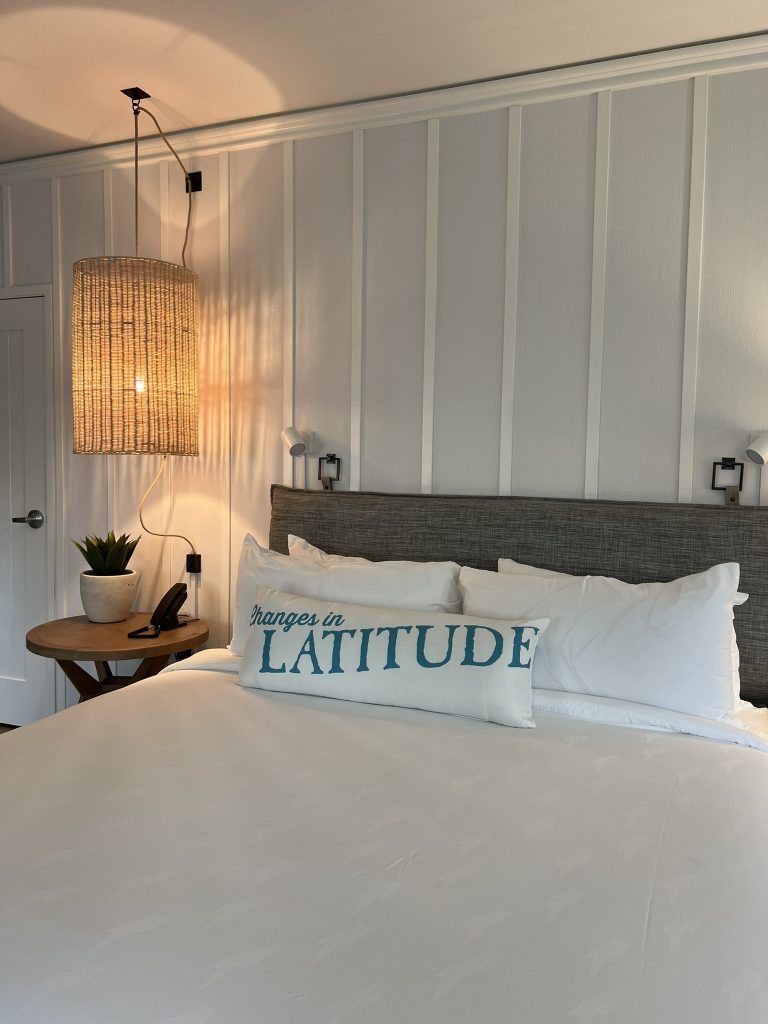 A cozy bed with a gray upholstered headboard, white bedding, and a decorative pillow reading "Changes in Latitude." A round wooden side table with a plant and telephone is beside the bed, under a woven pendant light.