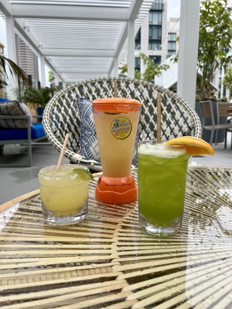 Three colorful cocktails on a glass-topped wicker table in an outdoor seating area.