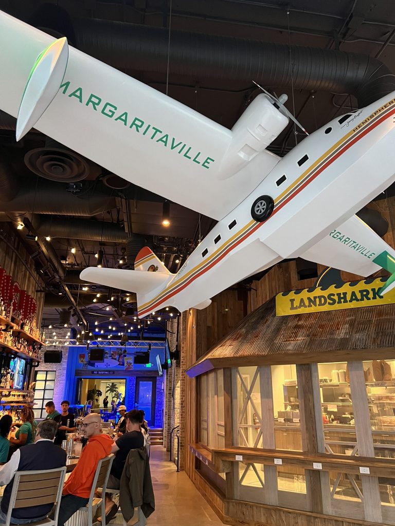 Interior of a restaurant featuring a large model airplane labeled "Margaritaville" hanging from the ceiling, with patrons seated at tables below.
