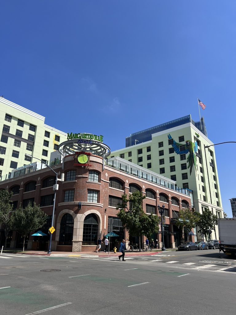 A city street scene with a multi-story hotel building that has a sign reading "Margaritaville" and features a large parrot mural. An American flag is visible, and there are people walking along the sidewalk.
