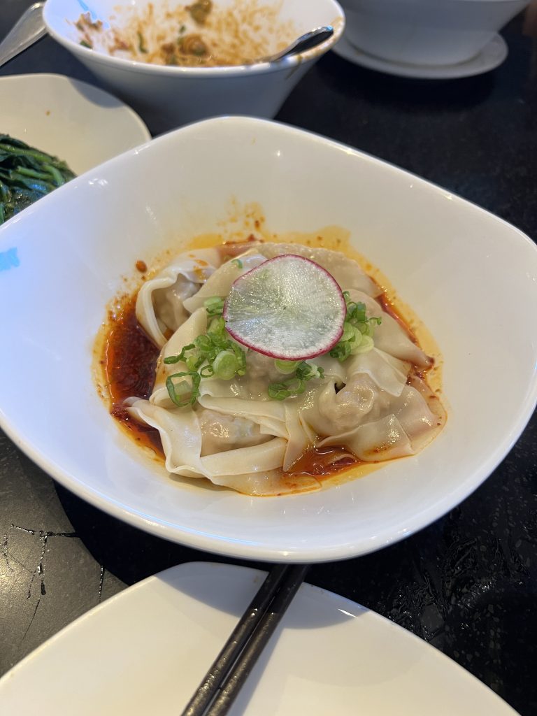 A dish of dumplings in chili oil garnished with sliced radish and green onions in a white bowl.