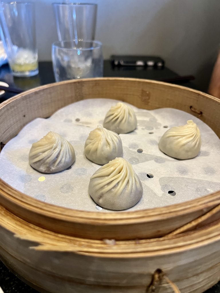 Steamed dumplings in a bamboo basket on a table with glasses in the background.