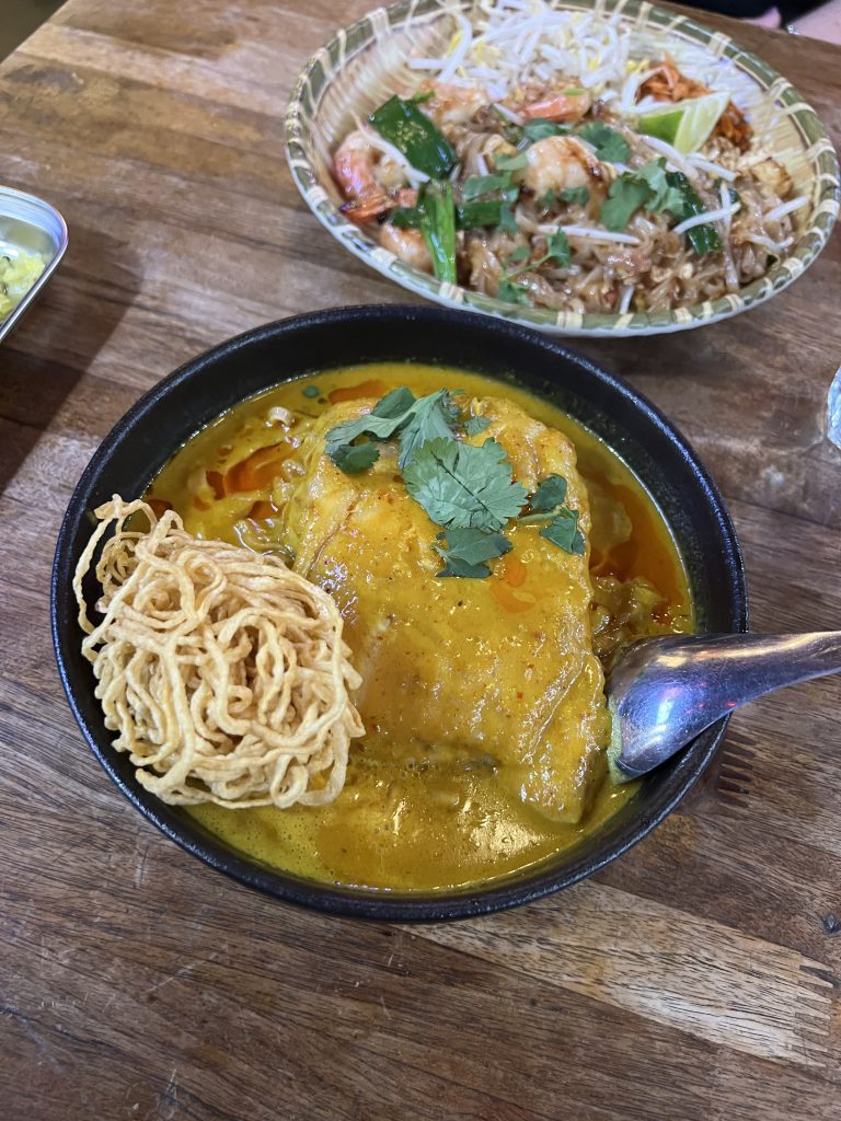 A bowl of curry with a piece of chicken, garnished with cilantro and crispy noodles, placed on a wooden table. Another dish with shrimp and noodles is visible in the background.