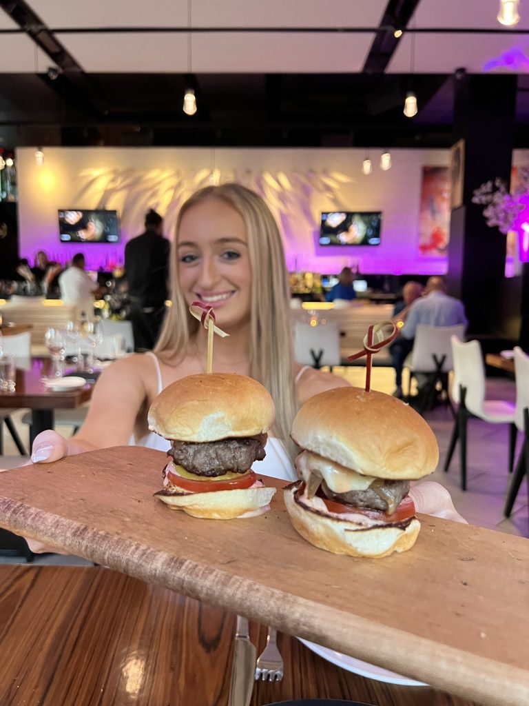 Person holding a wooden board with two small burgers in a restaurant setting.