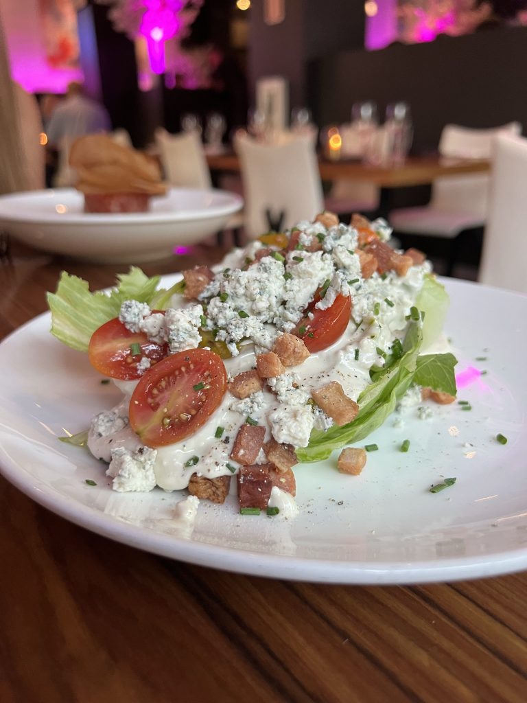 Wedge salad with iceberg lettuce, crumbled blue cheese, cherry tomatoes, bacon bits, and chives on a white plate.