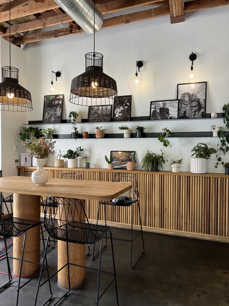 A modern cafe interior with a wooden table, wired chairs, and industrial-style hanging lights. The wall features black shelves displaying potted plants and black-and-white photographs. The ceiling has exposed wooden beams and ductwork.