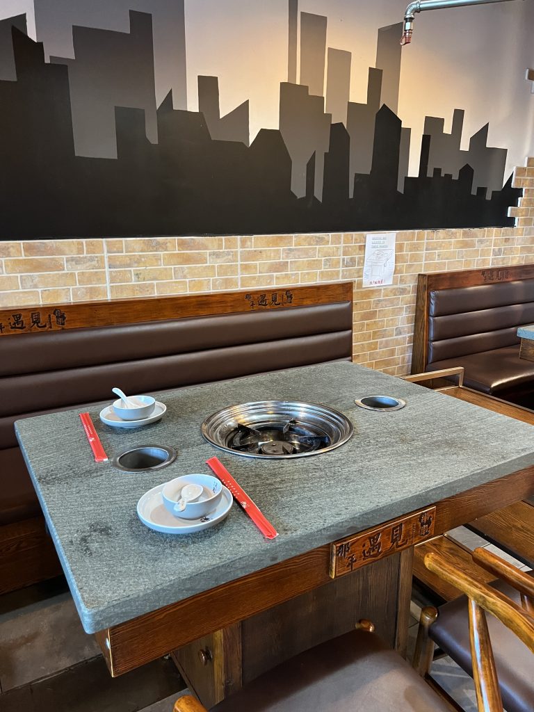 A restaurant table with a built-in grill, surrounded by wooden chairs. Tableware and red chopsticks are set on the surface. The background features a brick wall with a city skyline mural.