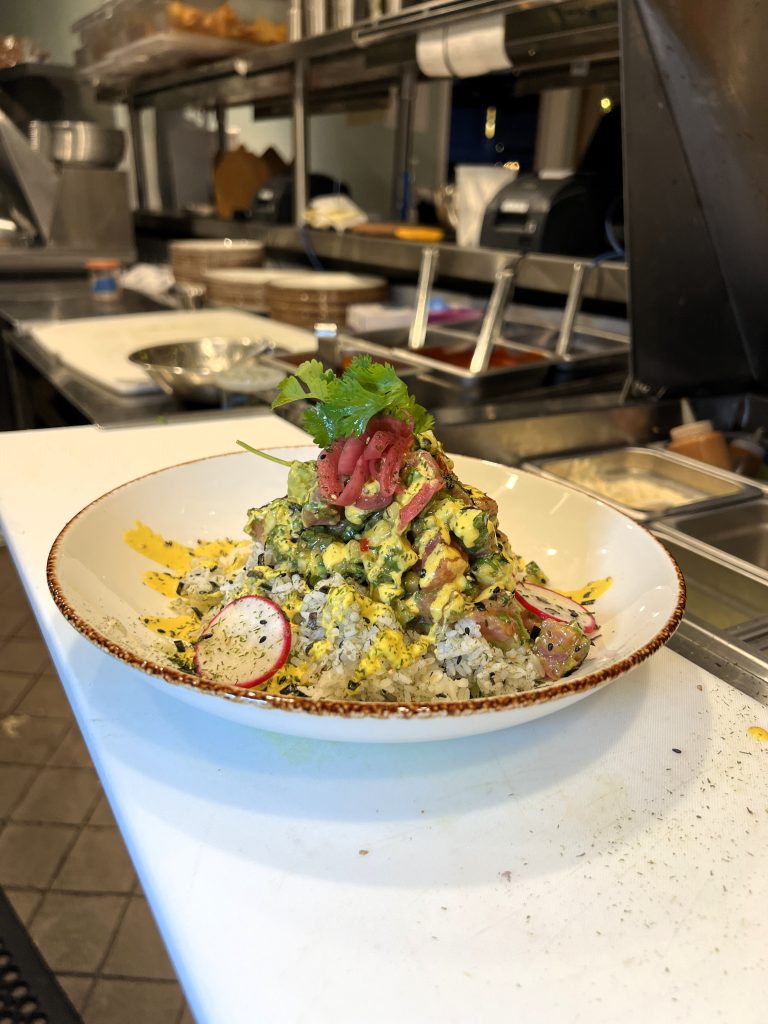 A plated dish of rice topped with a colorful salad, garnished with sliced radishes and fresh herbs, in a restaurant kitchen setting.