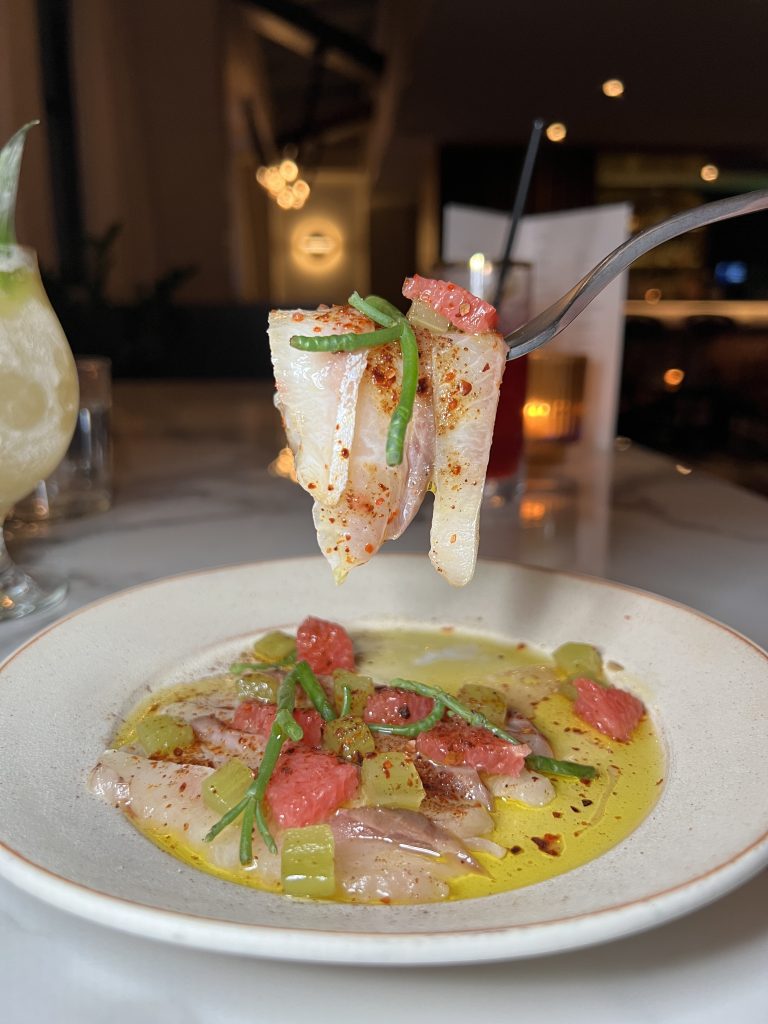 A fork holds a slice of sashimi garnished with small herbs and red seasoning, hovering above a plate of similar sashimi pieces, diced grapefruit, and cucumber in a light oil dressing.