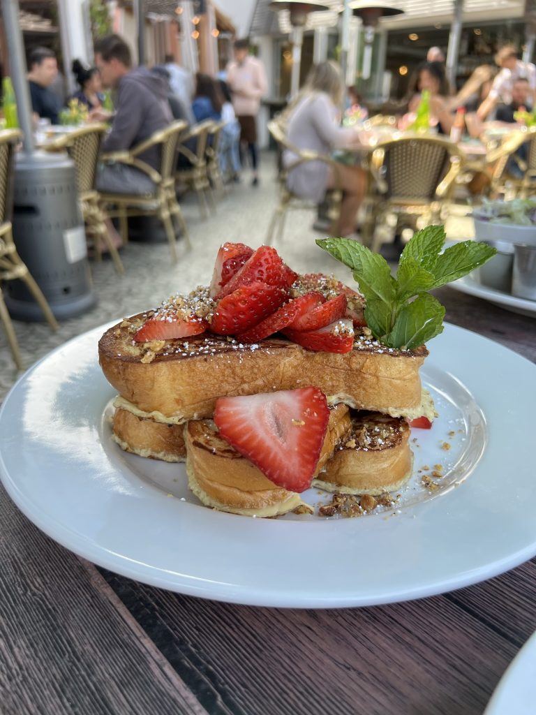French toast topped with sliced strawberries and a mint garnish on a white plate in an outdoor dining setting.