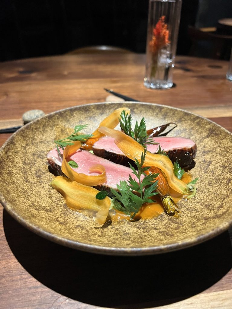 A plate with sliced pink meat garnished with thin carrot strips and herbs on a brown ceramic dish.
