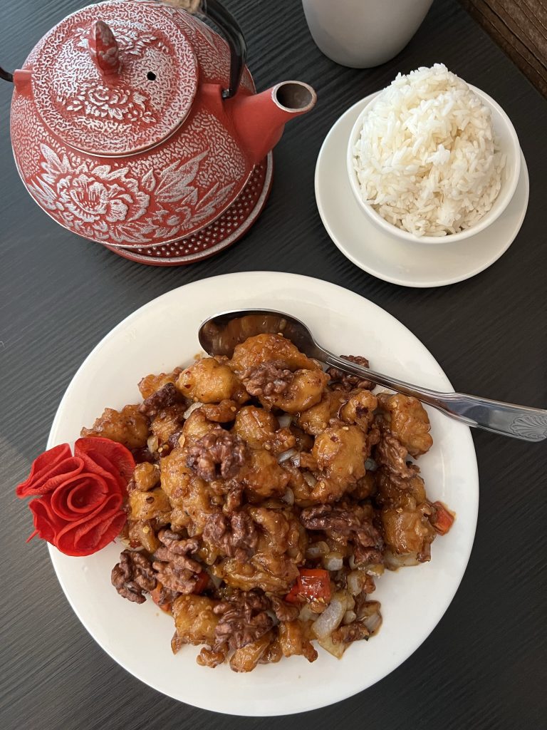 A plate of glazed walnut shrimp with a decorative red flower garnish, served alongside a bowl of white rice and a red patterned teapot.
