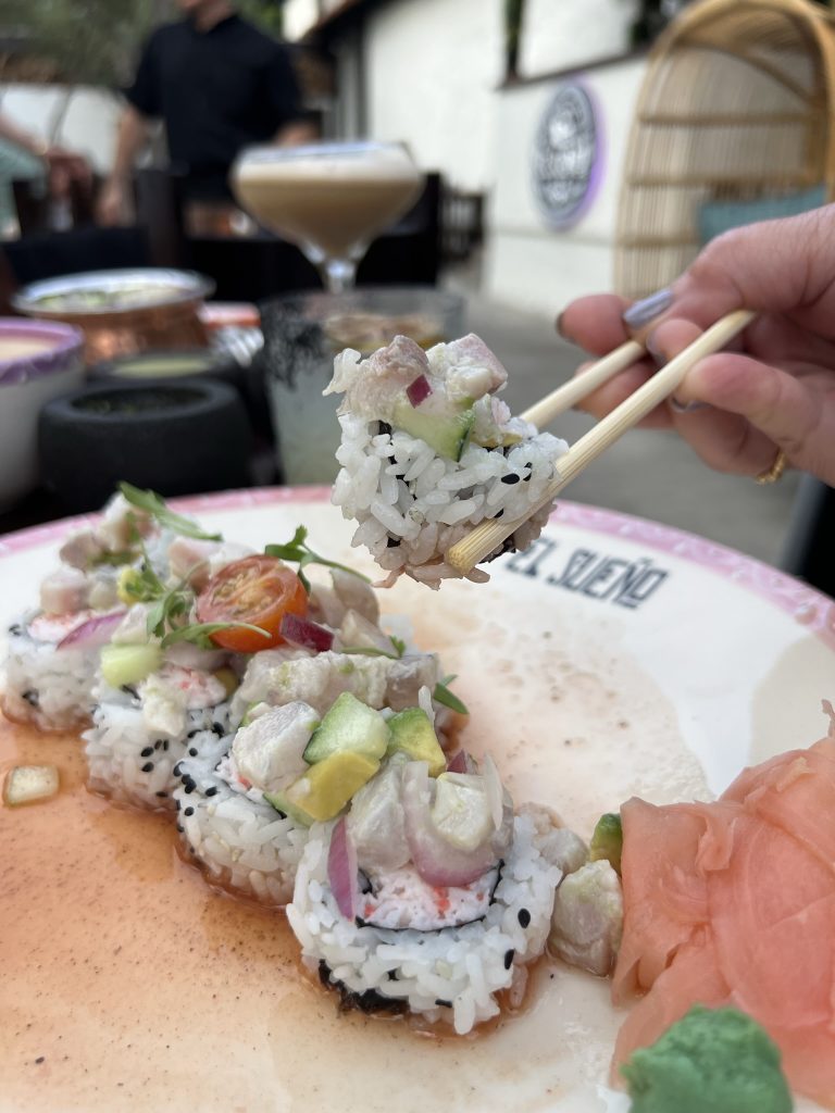 Sushi roll topped with diced avocado and fish, garnished with cherry tomato slices, on a plate with ginger and wasabi, being held with chopsticks.