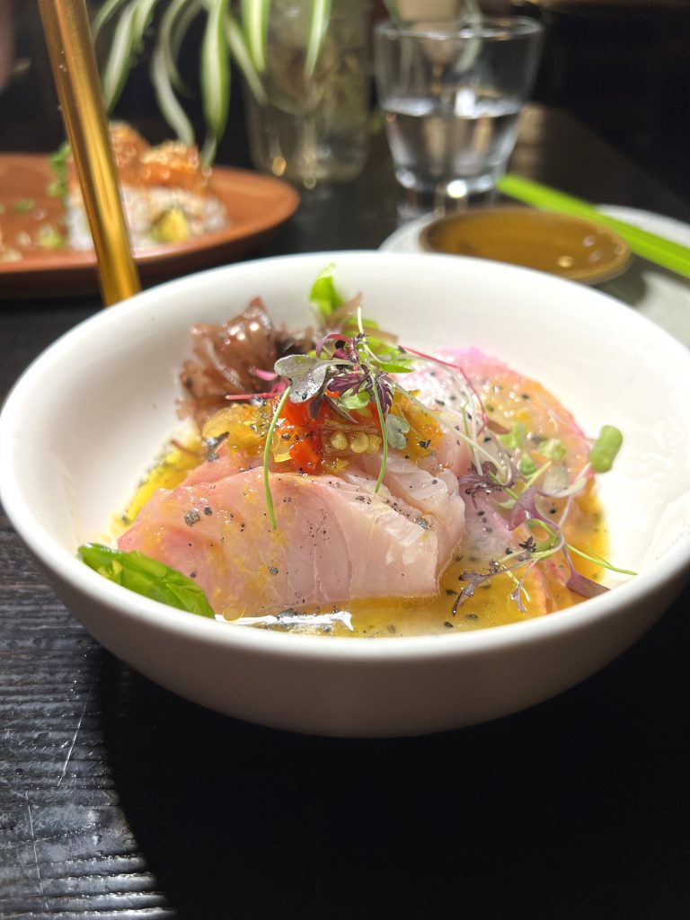 A bowl of colorful sashimi with microgreens and sauce garnished delicately in a white dish, on a dark table setting.