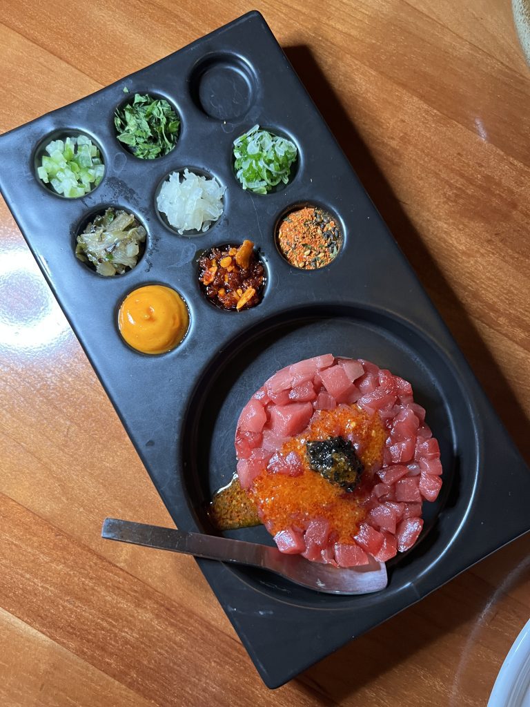 A dish of diced raw tuna garnished with red and black roe, accompanied by various herbs and sauces displayed in separate compartments on a black tray.
