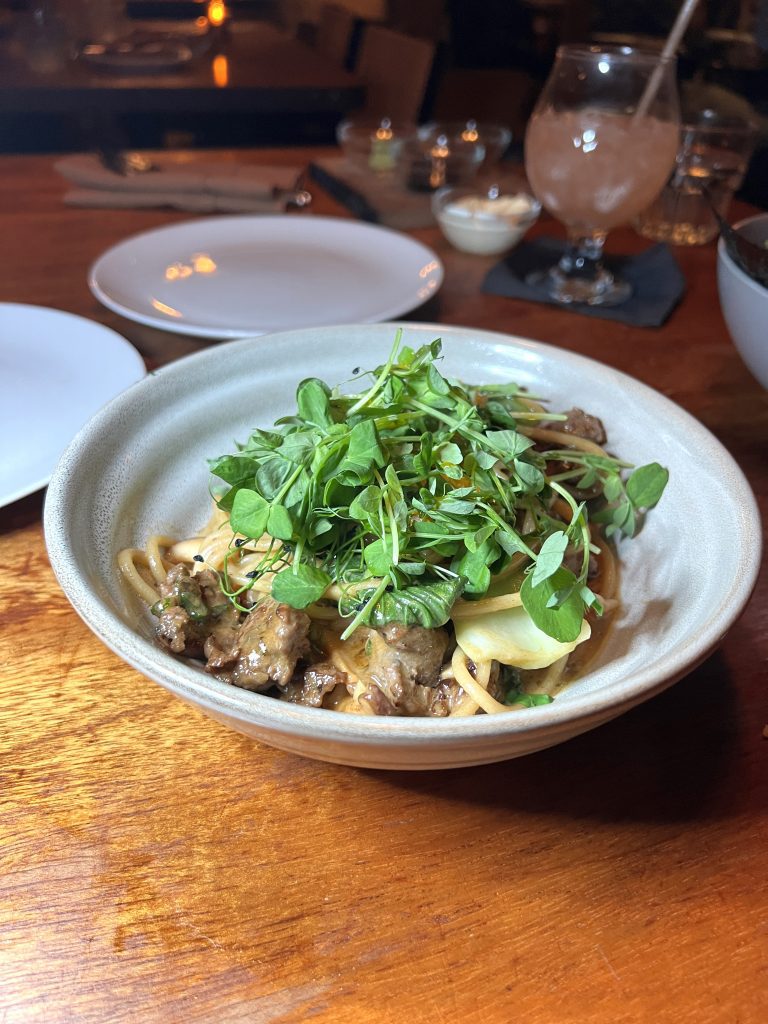 A bowl of pasta with ground meat and topped with a fresh green garnish, placed on a wooden table.