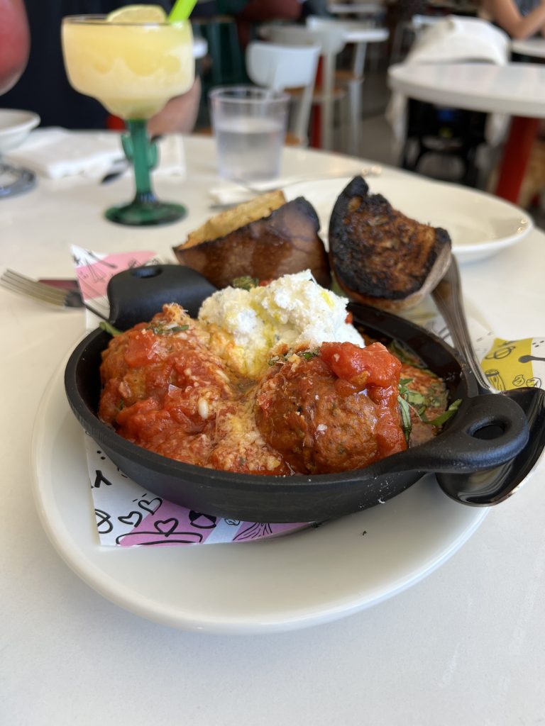 A skillet with meatballs in tomato sauce, ricotta cheese, and slices of charred bread, with a glass of frozen drink in the background.