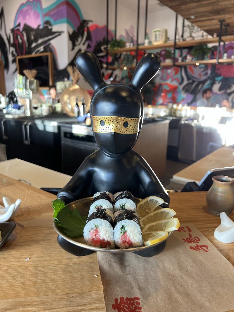 A black rabbit-shaped figurine holding a plate of sushi rolls with lemon slices, placed on a wooden counter in a restaurant with colorful graffiti on the walls.