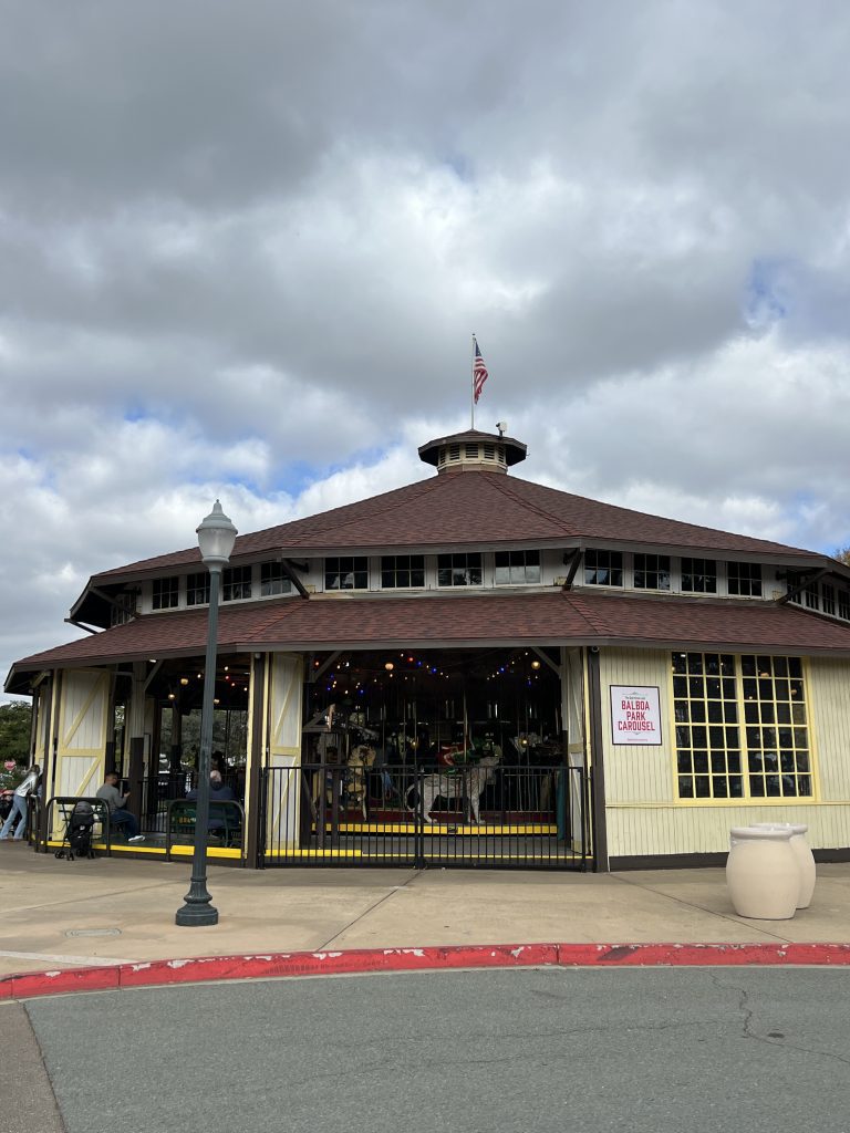 Circular building with a carousel inside, labeled "Balboa Park Carousel," with a cloudy sky and an American flag on top.
