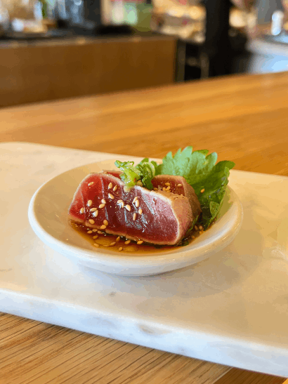 A piece of seared tuna garnished with sesame seeds and cilantro on a small plate with soy sauce.