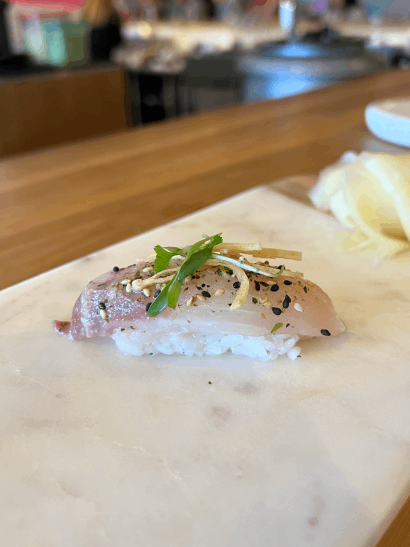 A piece of sushi topped with fish, garnished with herbs and sesame seeds, on a marble surface.