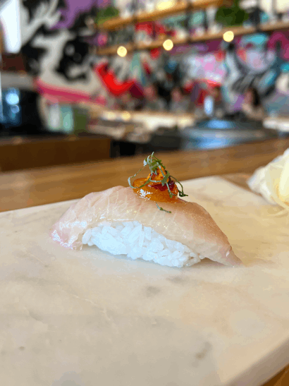 A piece of sushi on a marble slab topped with fish, garnished with herbs and orange sauce.