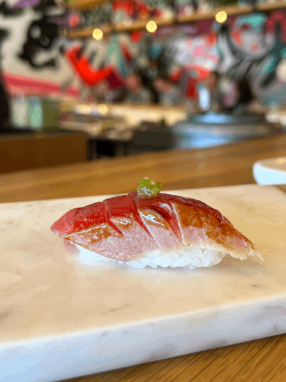 A piece of sushi with a slice of fish on top, garnished with a small amount of green seasoning, placed on a marble surface in a vibrant restaurant setting.