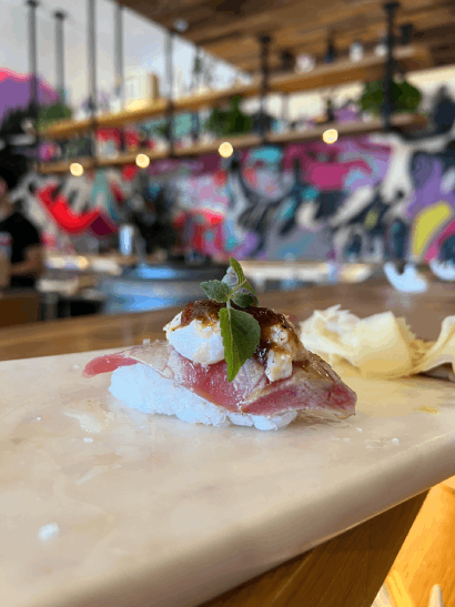 Sushi piece garnished with herbs on a marble surface, with a blurred colorful interior backdrop.
