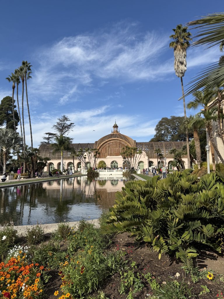 A historical botanical building with arches reflected in a pond, surrounded by lush greenery and flower beds under a blue sky.