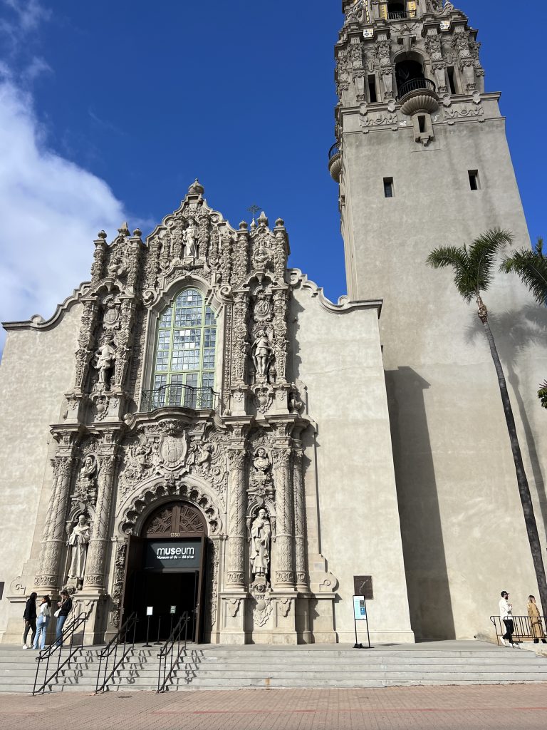 Historic building with ornate stone carvings, featuring an arched entrance labeled "museum" and a tall tower, under a clear blue sky.