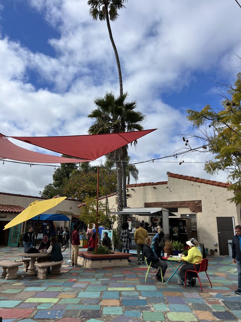 Outdoor courtyard with colorful tile flooring, people seated at tables, and others gathered around a small building under shade sails with palm trees and a cloudy sky above.
