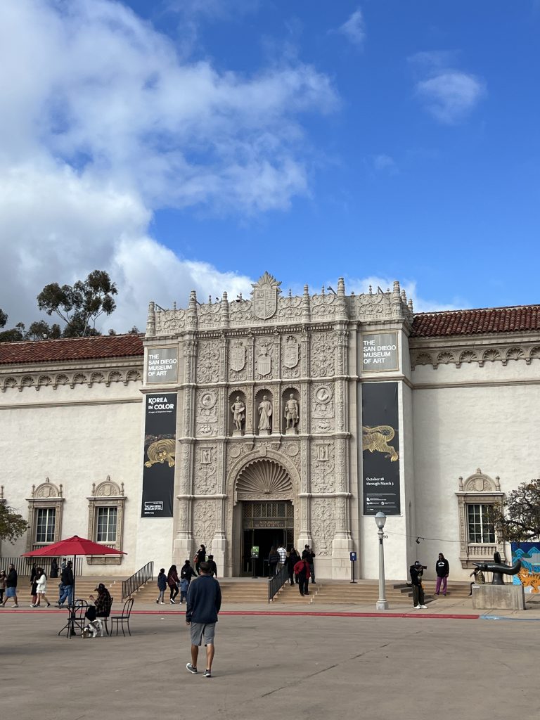 The San Diego Museum of Art building facade with intricate architectural details and banners, people walking nearby.