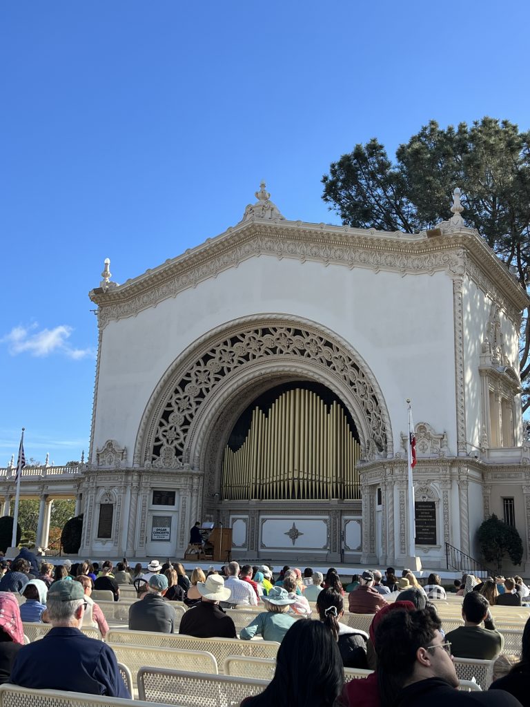 People seated in an outdoor amphitheater in front of a large ornate organ pavilion under a clear blue sky.