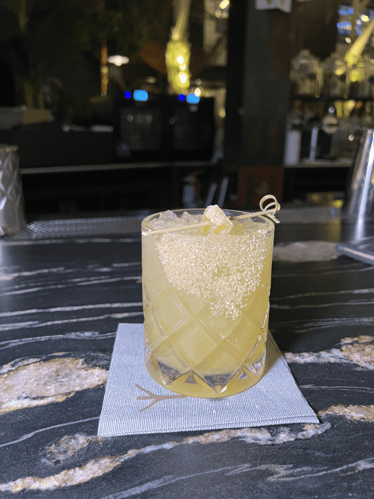 A yellow cocktail in a textured glass on a napkin, garnished with sugar and a candied ginger slice, set on a dark marble countertop.