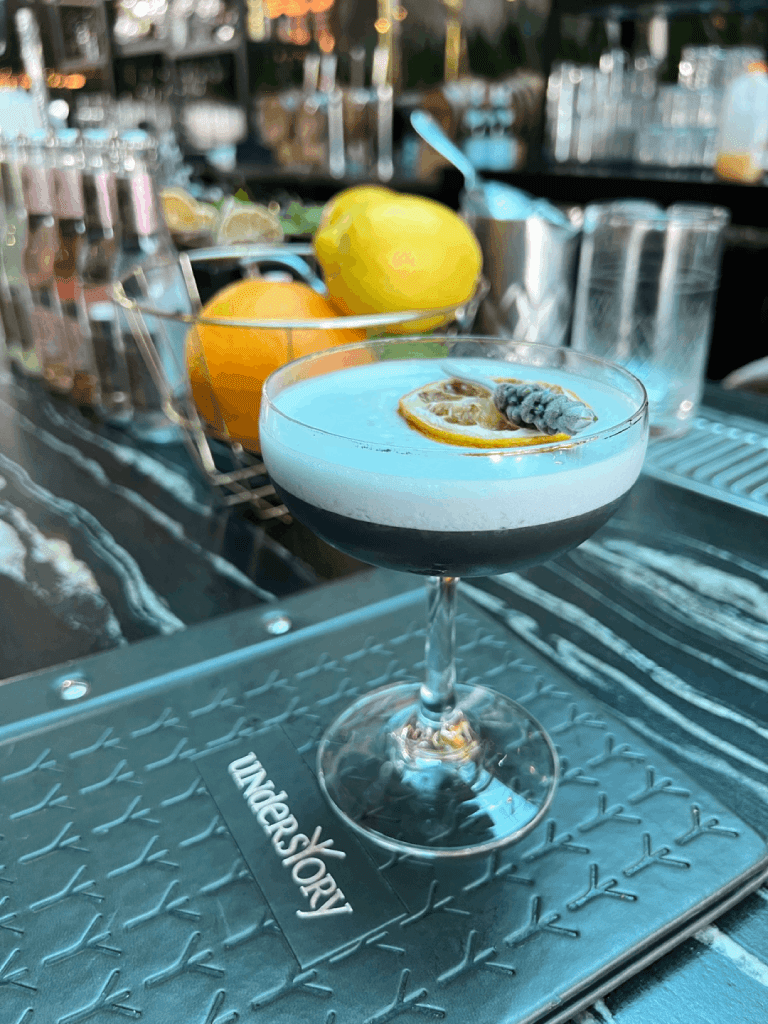A cocktail in a coupe glass garnished with a dried orange slice and a sprig of herbs, placed on a bar counter with fruit and bottles in the background.