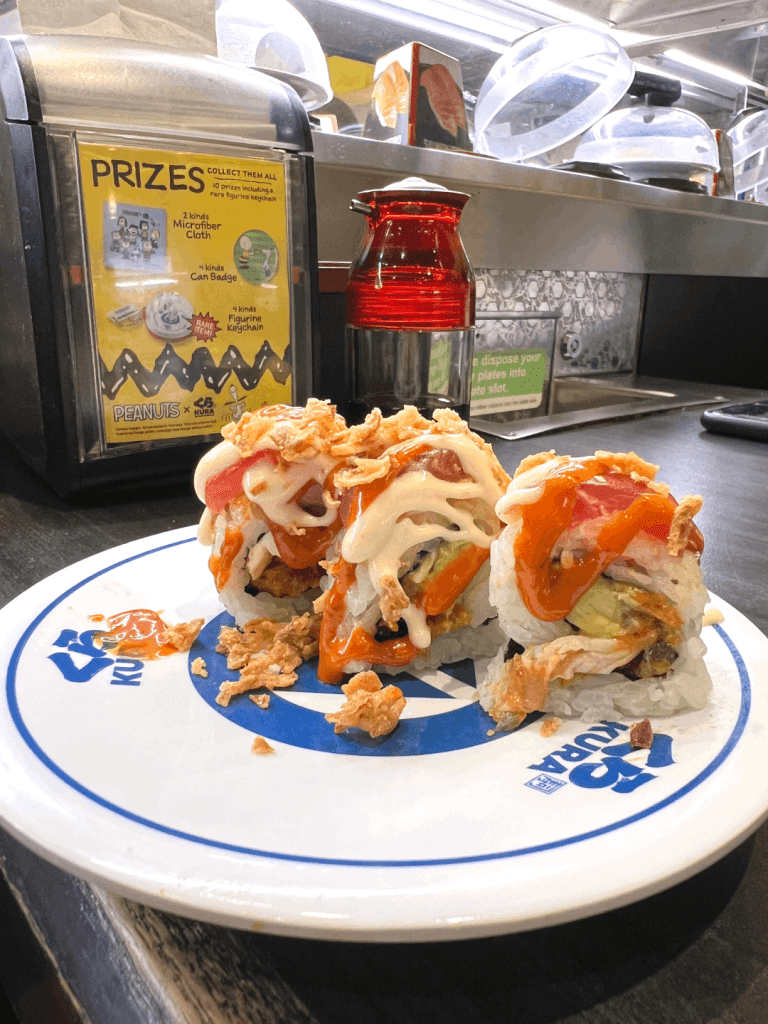 Sushi rolls topped with sauce and crispy onions on a Kura sushi plate, with a red soy sauce dispenser and a prize display in the background.