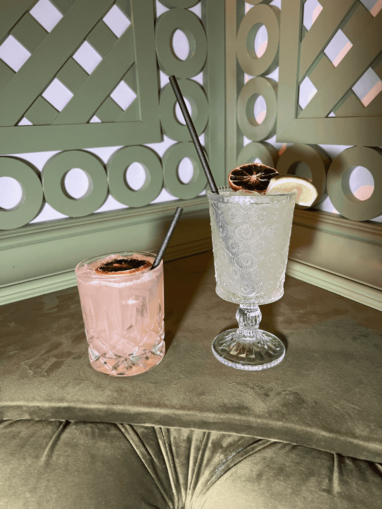 Two cocktails on a velvet surface, one in a crystal glass with a pink drink and dried lime, and the other in an ornate glass with a clear drink, lemon slice, and dried lime garnish.